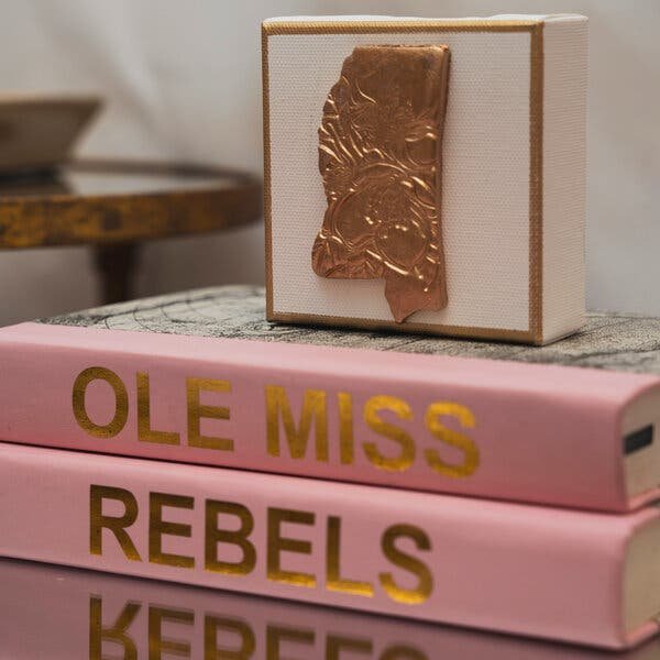 A decorative box sits atop a stack of pink-covered books, with the top book featuring 'Ole Miss' in gold lettering on the spine